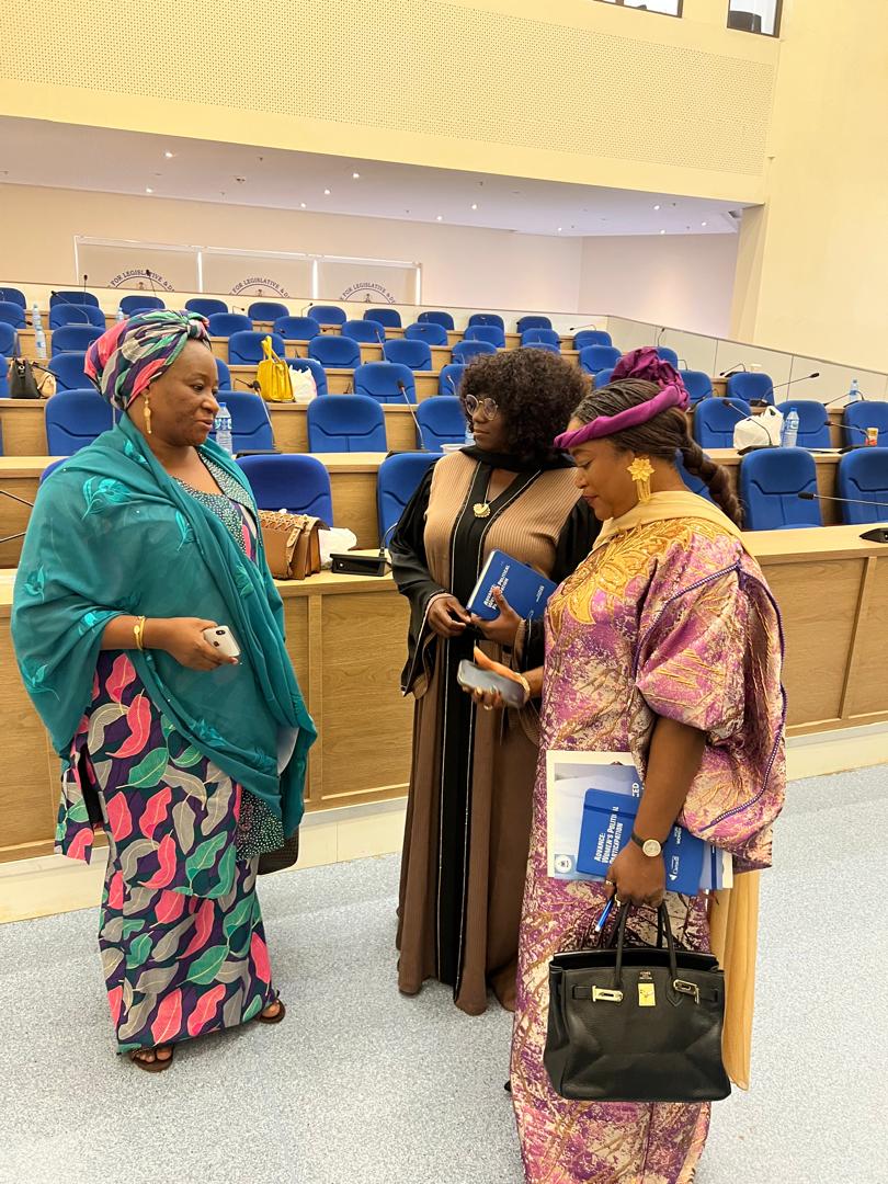 Three women in conversation inside a legislative chamber between sessions.